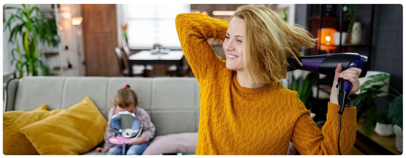 Frau die ihre Haare föhnt. Im Hintergrund sitzt ein Kind.